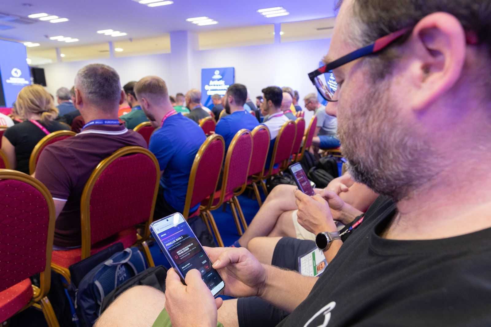 A judge entering a score on his phone, the full hall of judges behind him.